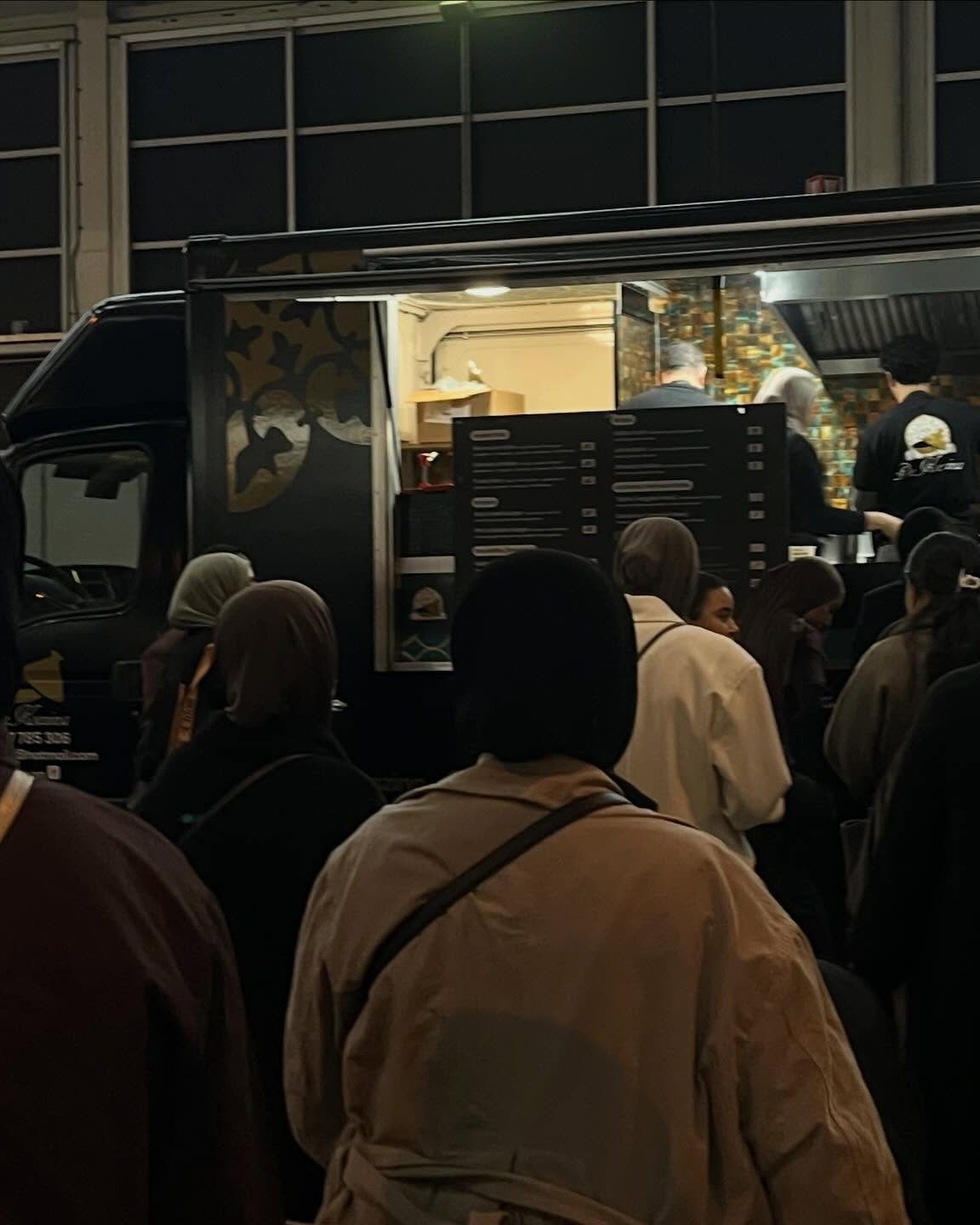 De zwarte De Koezina-foodtruck met open luik en warm licht, gasten in de rij